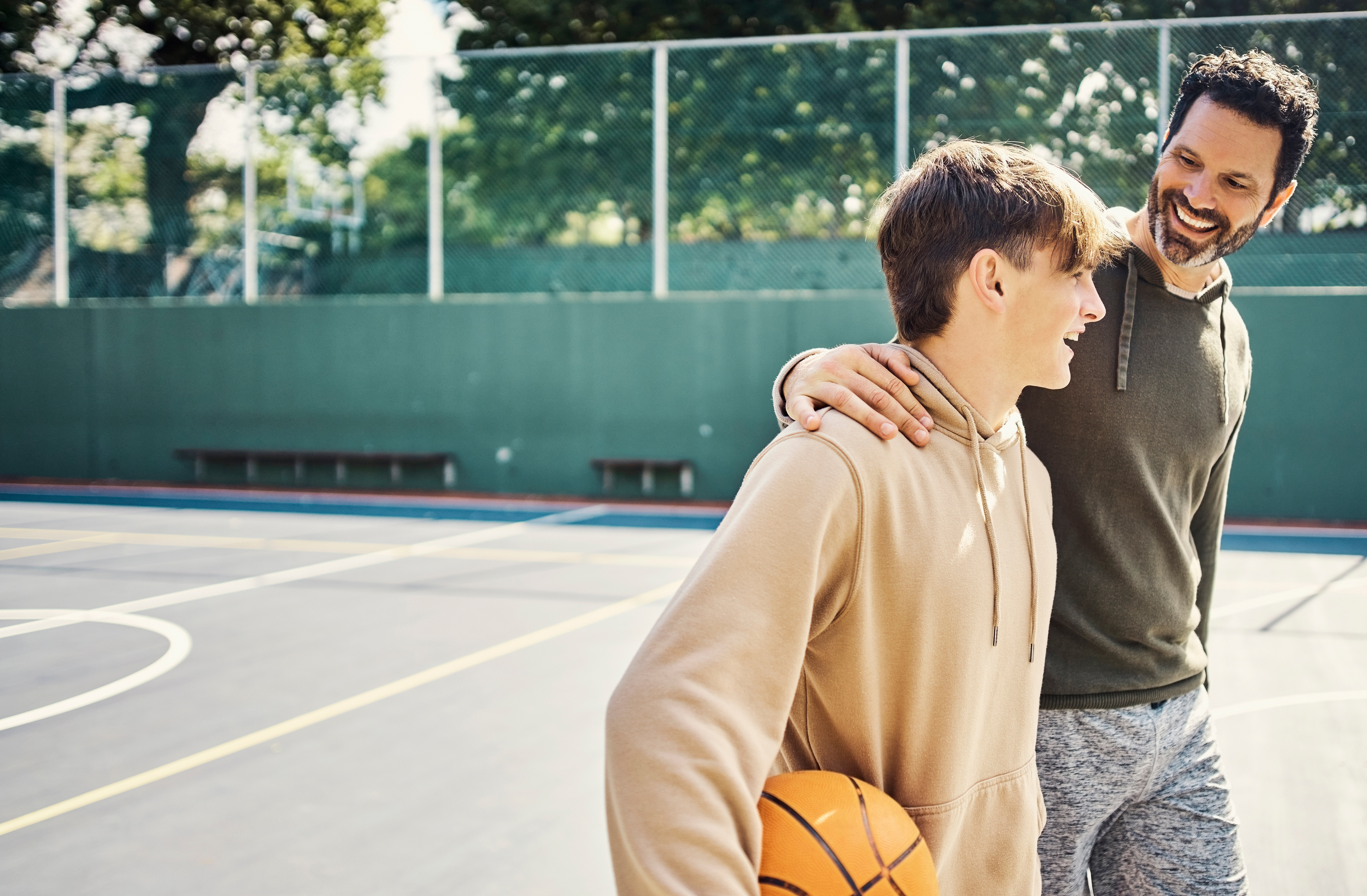 Een jongen loopt op een basketbalveld met een basketbal onder zijn arm en een jongerenwerker die zijn arm om zijn schouder heeft geslagen
