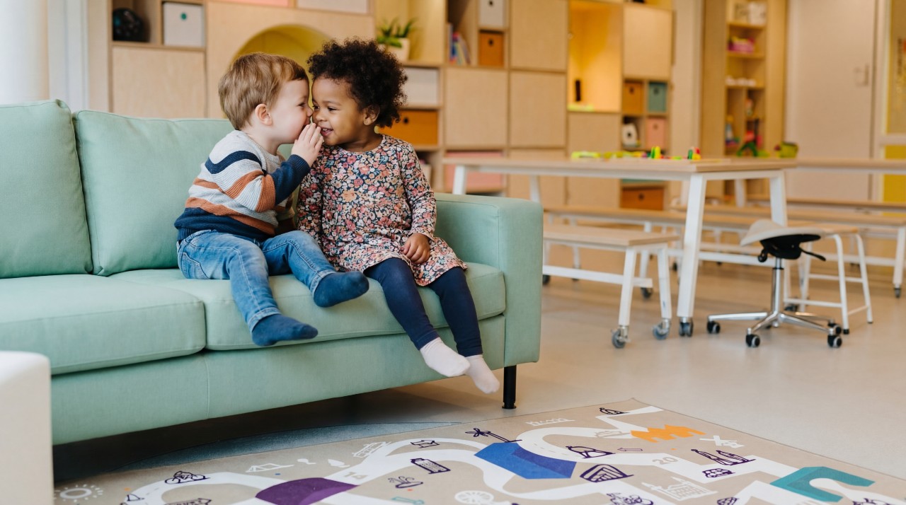 Twee peuters, een jongen en een meisje, fluisteren met elkaar op een bank op een peuteropvang van gro-up