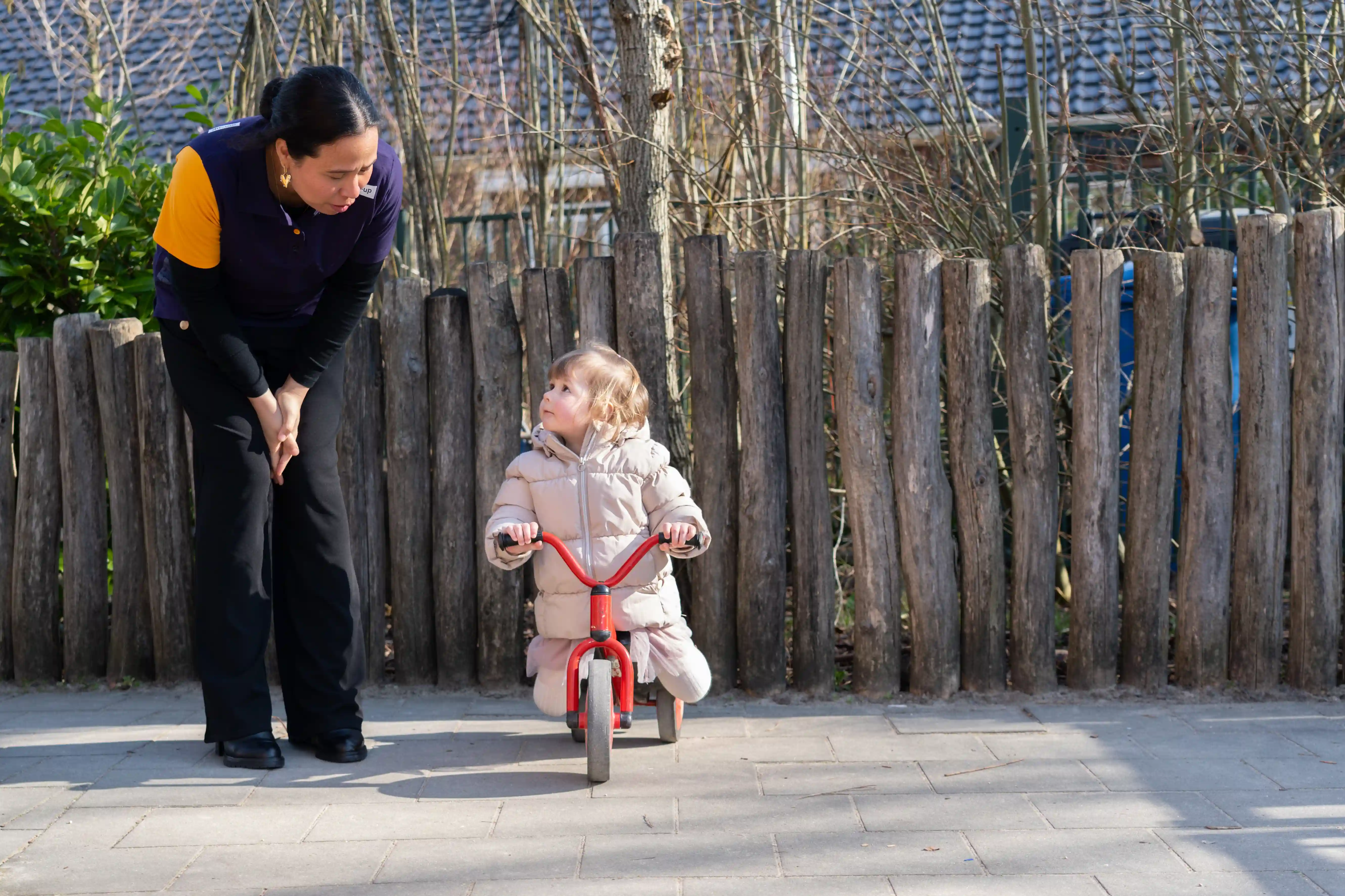 Een pedagogisch medewerker helpt een peuter meisje die wil fietsen op een driewieler buiten bij een peuteropvang van grro-up