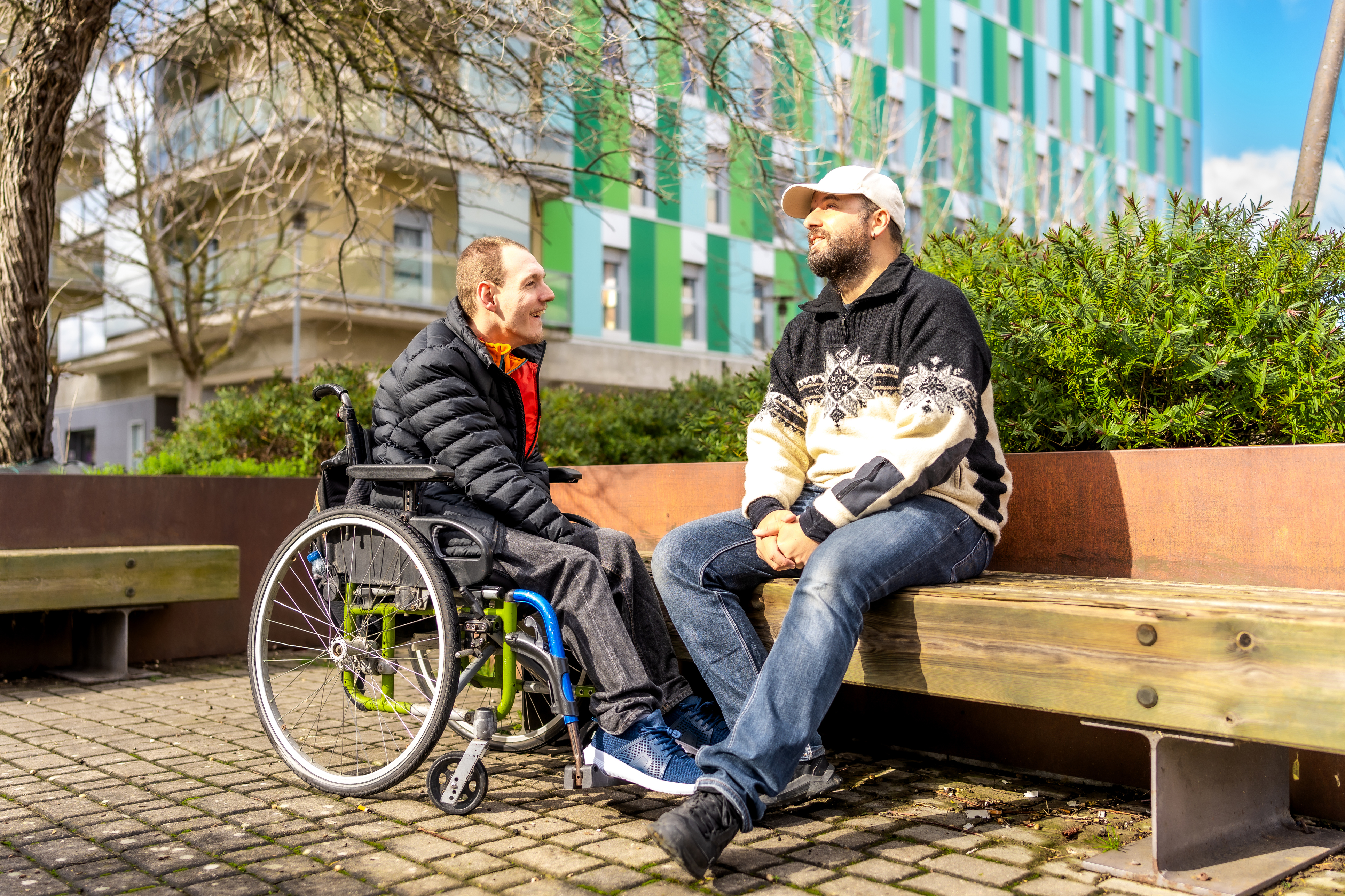 Twee mannen, een in een rolstoel en een op een bankje, zitten te praten over hun wijk