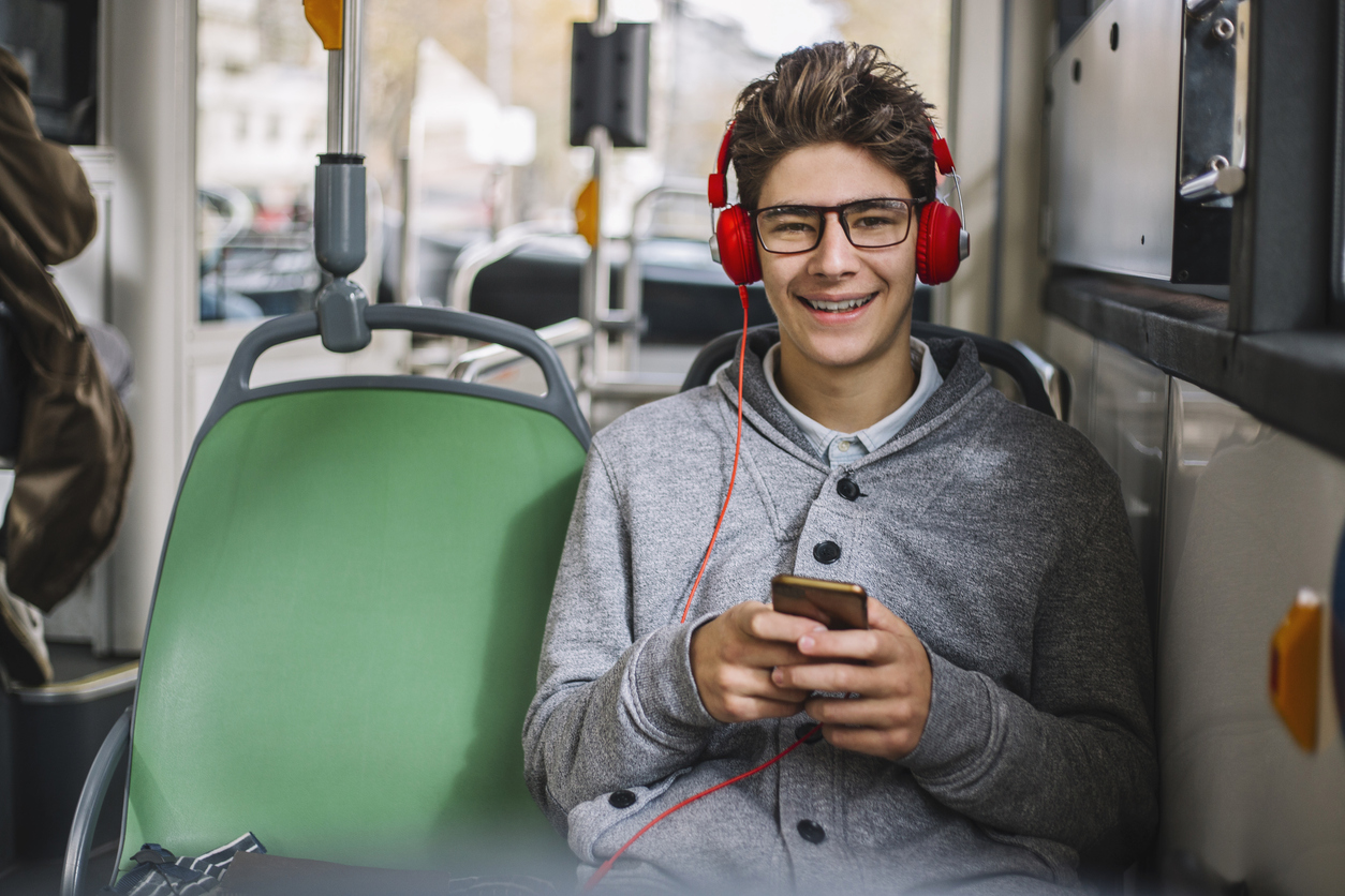 Tienerjongen lacht in de bus lacht in de camera. Hij heeft rode koptelefoon op zijn hoofd en een smartphone in zijn handen.