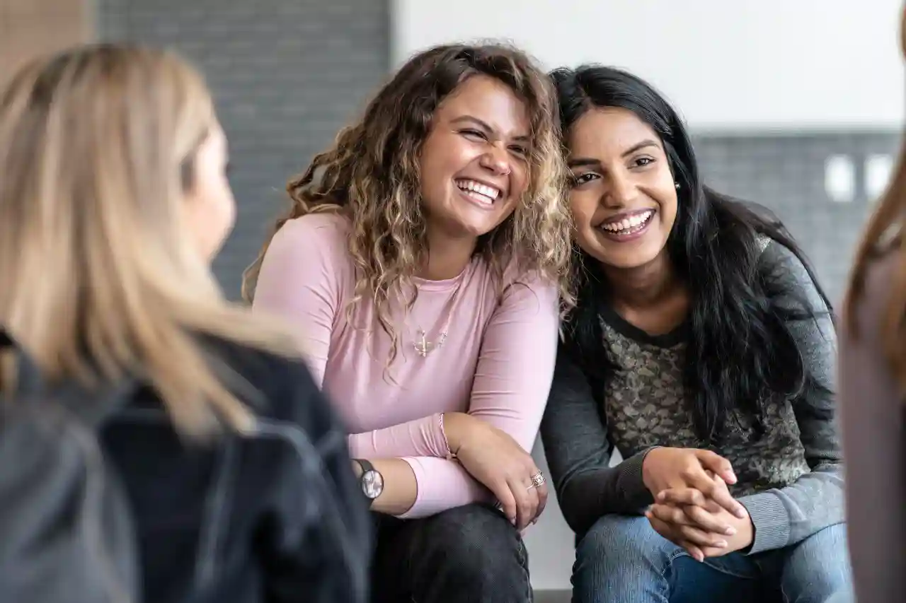 Twee tiener meiden lachen met de hoofden tegen elkaar na twee dames die met hun rug naar de camera zitten